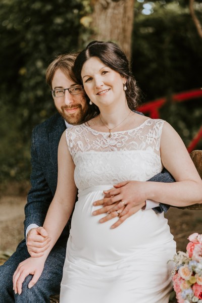 Bride and groom are seated she places hand on pregnant belly and on grooms hand