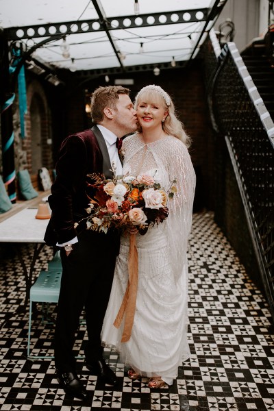 Groom kisses his bride wife on the cheek as she holds bouquet