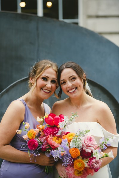 Bride and bridesmaid smile holding colourful bouquet flowers