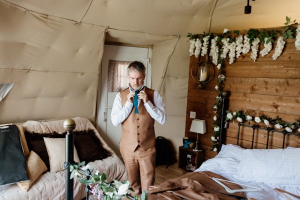 Groom gets ready in room buttoning shirt and fixing tie