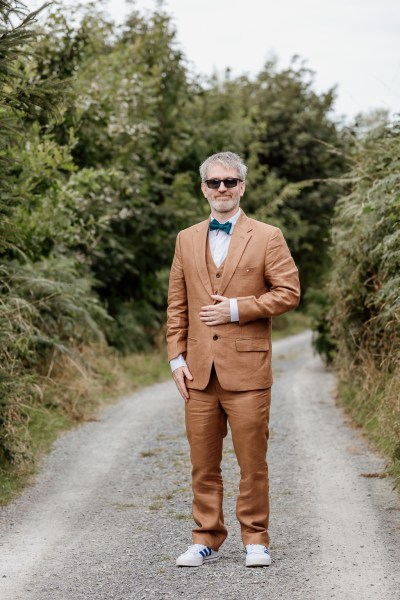 Groom in brown suit stands along pathway to park forest grounds wearing sunglasses