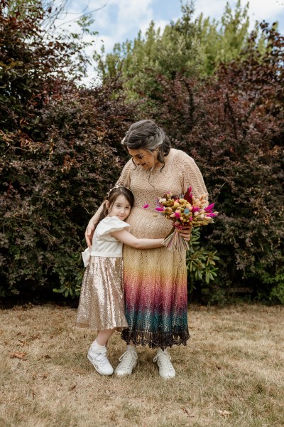 Bride and her daughter stand on the grass in forest setting daughter hugs her mum