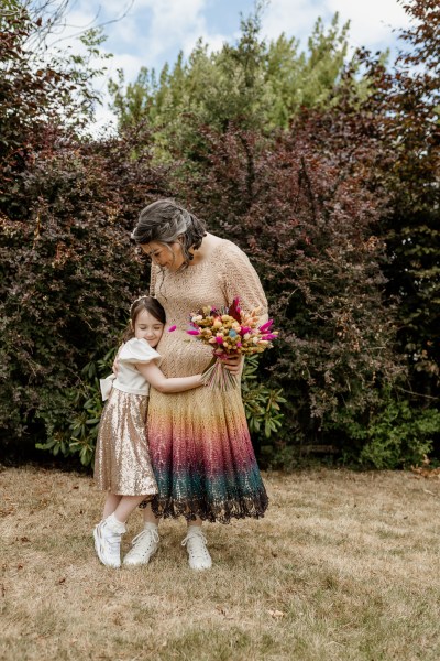 Bride and her daughter stand on the grass in forest setting daughter hugs her mum