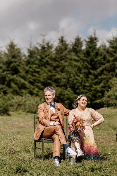 Bride and groom sit in farm forest trees behind them bouquet in shot