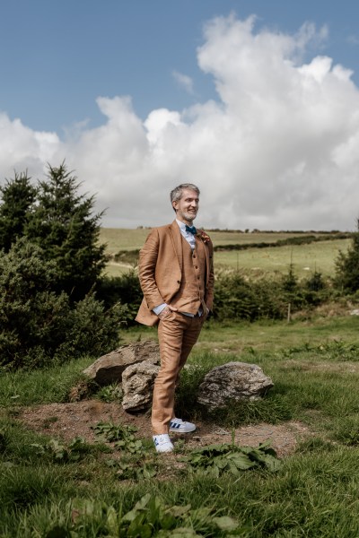 Groom stands in front of greenery scenery on the grass farm trees in background