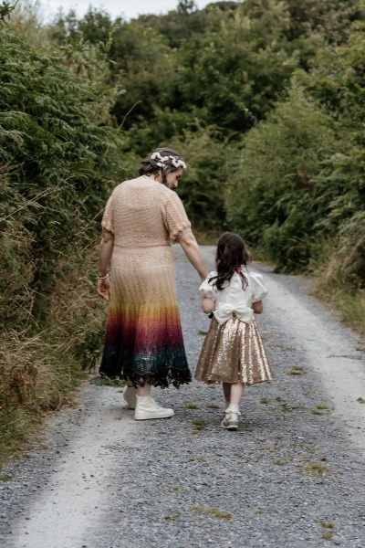 Bride holds daughters hand from behind shot as they walk the road/pathway