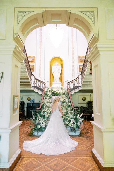 Bride stands on her own at bottom of staircase covered in flowers