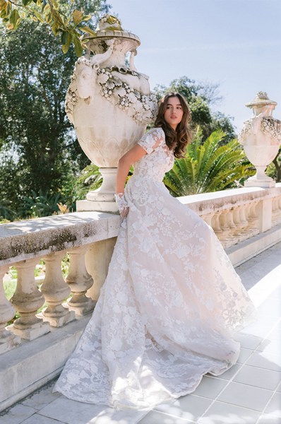 Monique Lhuillier Spring 2024 Collection bride leaning against pillar in garden bridal gown lace