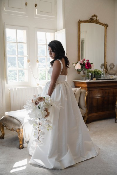 Bride gets ready stands by windowsill holding white bouquet flowers