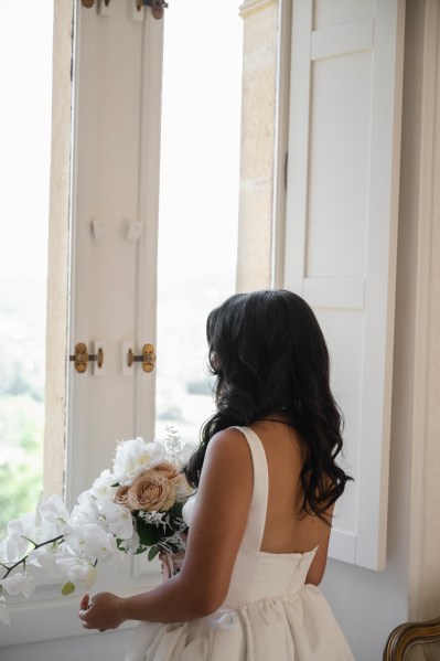 Bride gets ready stands by windowsill holding white bouquet flowers looks out of window