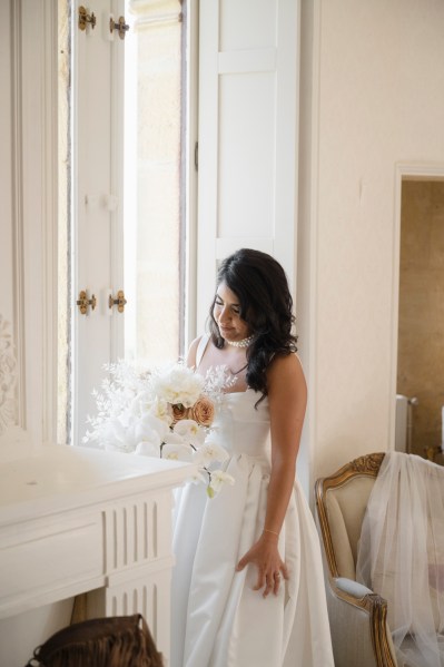 Bride gets ready stands by windowsill holding white bouquet flowers