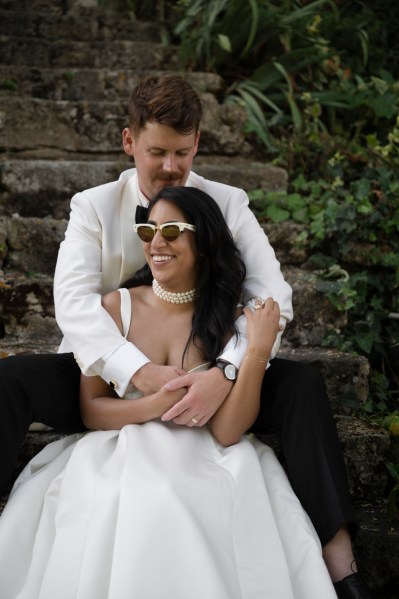 Couple wear sunglasses bride and groom sit amongst flowers on steps in forest smiling