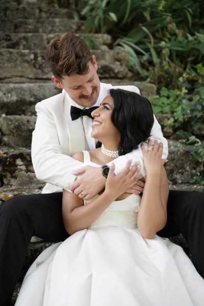 Bride smiles as groom smiles at her from above arms around her