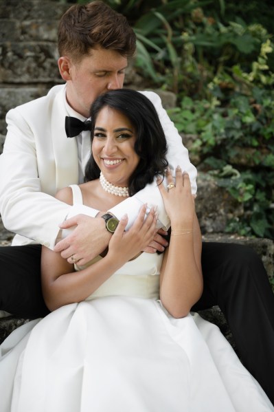 Bride smiles as groom kisses her on the head arms around her