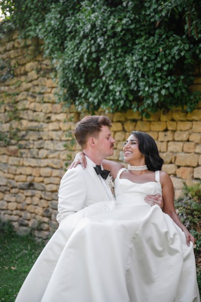 Groom picks up bride on grass bricked wall in background