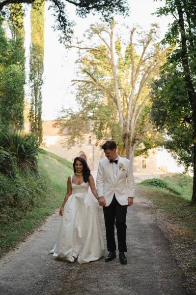 Groom looks at his bride as they walk along pathway to forest