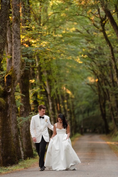 Bride and groom walk in forest surrounded by trees along pathway