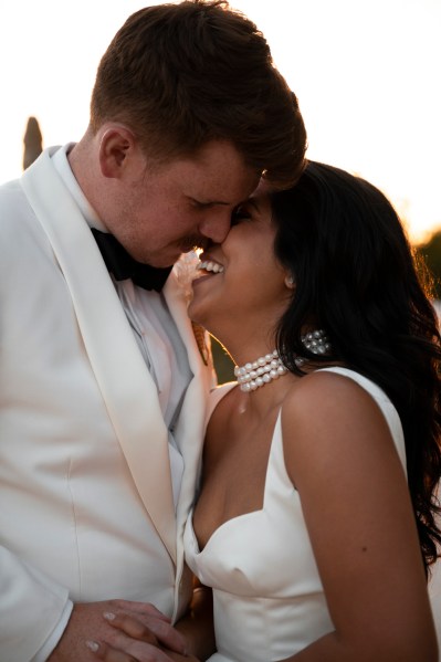 Bride and groom laugh as they hold each other sunset in background going in for a kiss and laughing