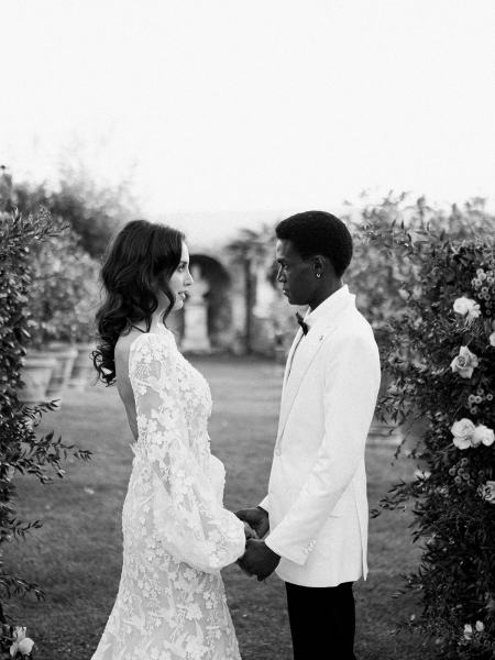 Bride and groom holding hands between hedges in garden black and white