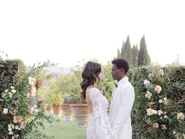 Bride and groom face each other standing hand in hand flowers in hedges either side of them