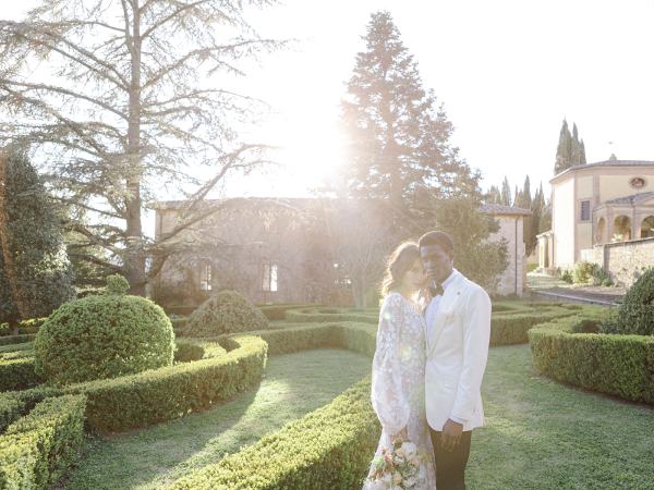 Bride stands with bouquet in hand with groom on the grass in garden wide shot