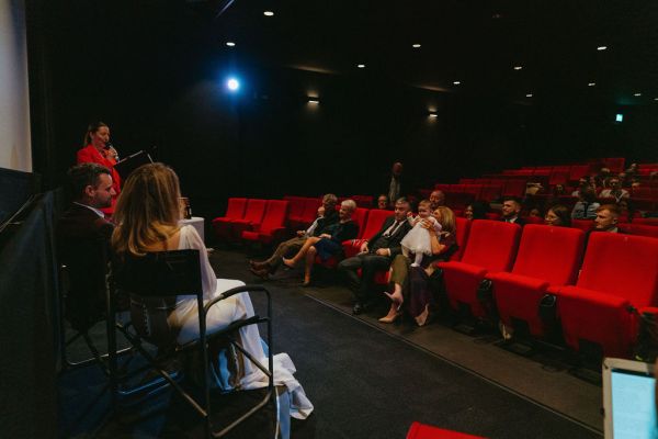 Bride and groom sit on stage alter in front of audience members sitting in red seats