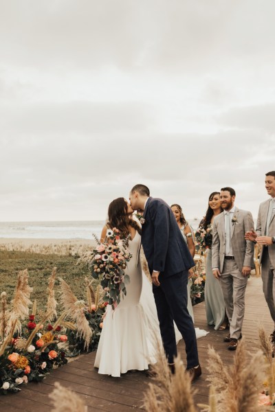 Bride and groom bridesmaids and groomsmen pose for a picture beach setting they kiss