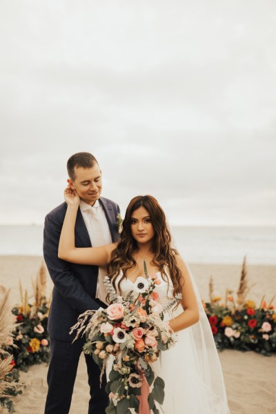 Bride hand on grooms cheek sea in background setting bouquet