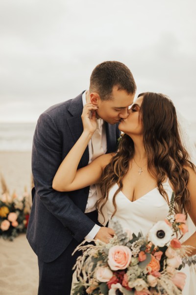 Bride has hand on grooms cheek beach setting they kiss