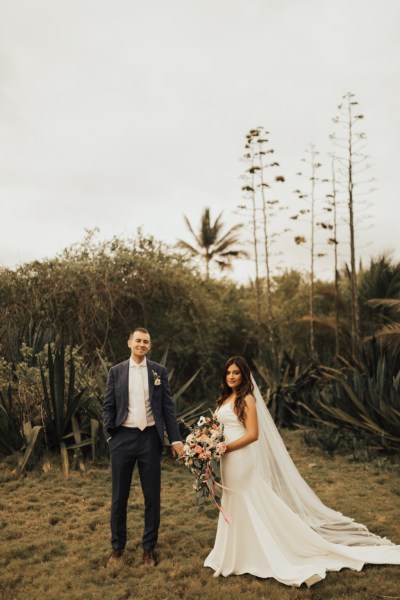 Bride and groom standing on grass walking in front of palm trees beach setting facing the camera