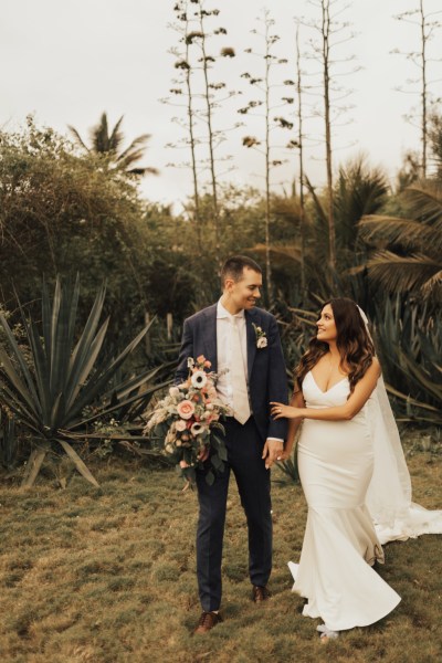 Bride and groom standing on grass walking in front of palm trees beach setting they look at each other