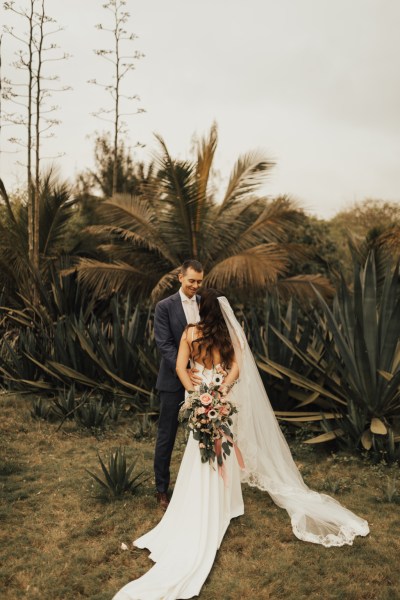 Bride and groom go in for a kiss palm trees in background bouquet behind brides back