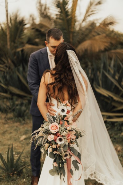 Bride and groom go in for a kiss palm trees in background bouquet behind brides back