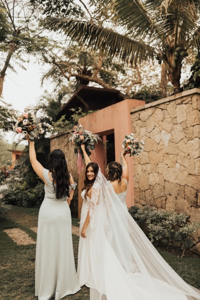 Bride and her bridesmaids standing with bouquet/flowers garden setting from behind flowers in the air bride looks over her shoulder