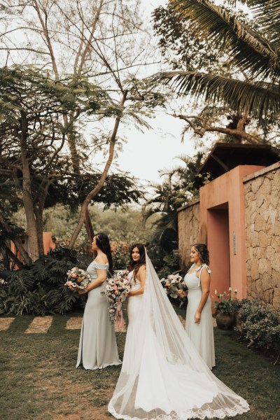 Bride and her bridesmaids standing with bouquet/flowers garden setting from behind flowers in the air bride looks over her shoulder