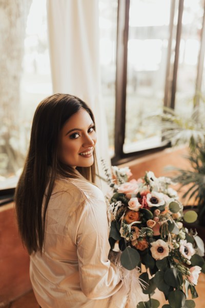 Bride looks over her shoulder holding bouquet at window