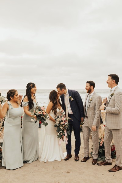 Bride and groom bridesmaids and groomsmen pose for a picture beach setting they kiss