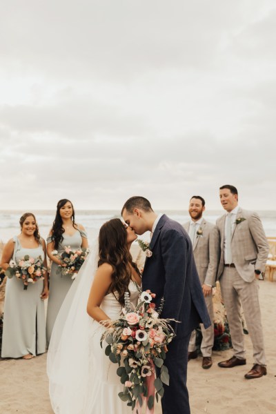 Bride and groom bridesmaids and groomsmen pose for a picture beach setting they kiss