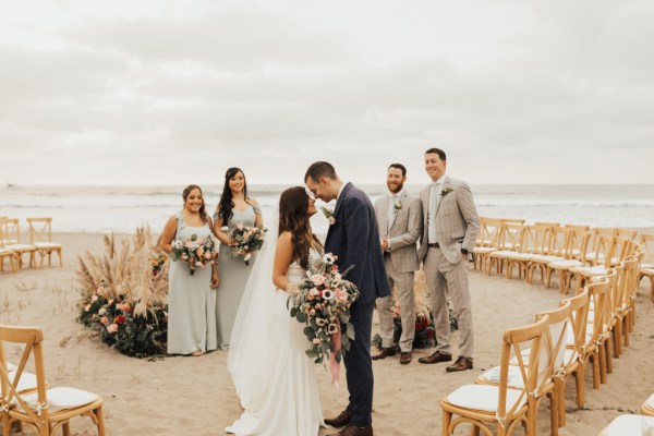 Bride and groom bridesmaids and groomsmen pose for a picture beach setting they kiss