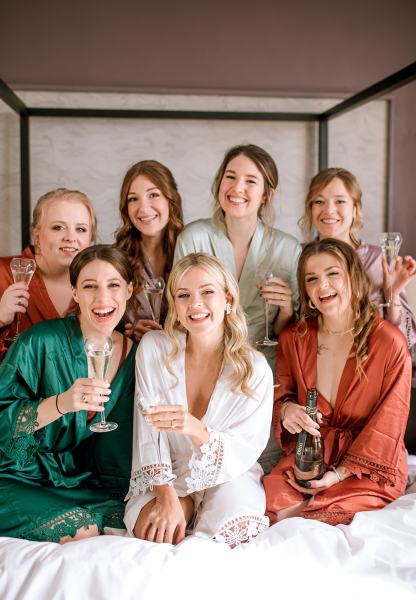 Bride bridesmaids on bed holding out glasses of champagne on bed smiling