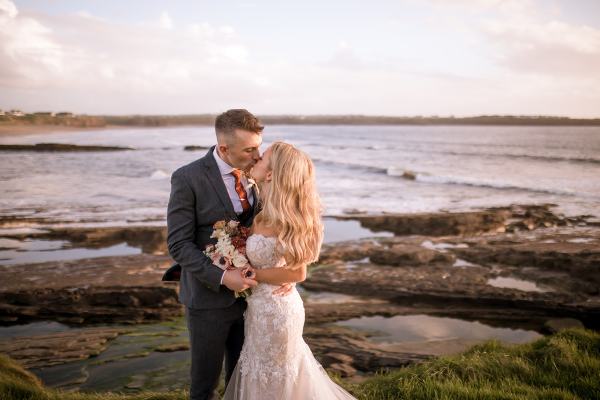 Groom has arm around brides waist they laugh beach setting they kiss