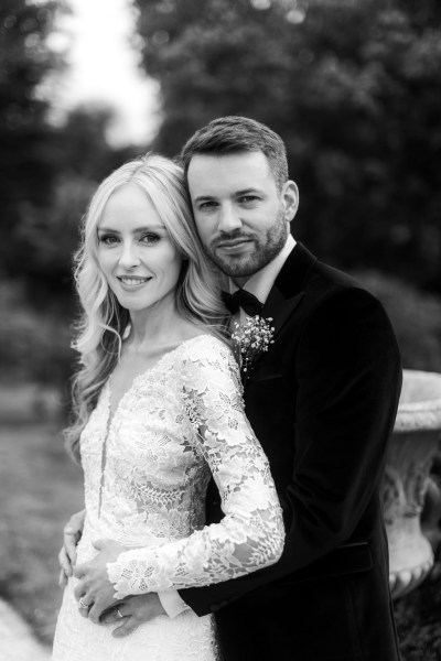 Black and white groom has arms around brides waist they face the camera