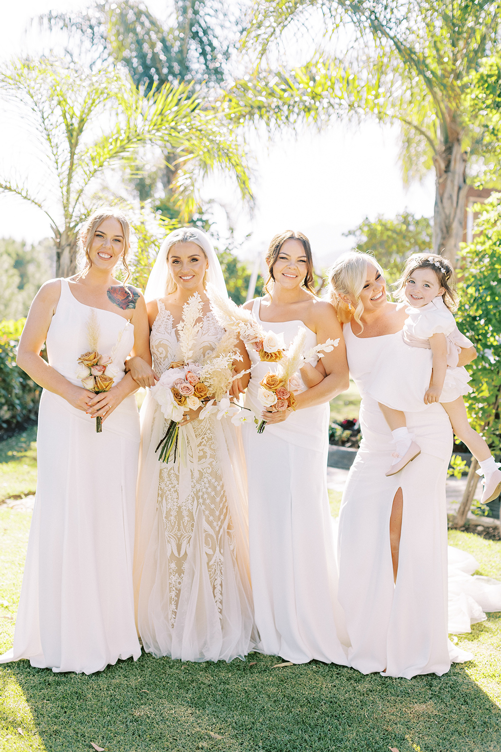 Bride and her bridesmaids stand and pose on the grass garden setting with little girl in shot