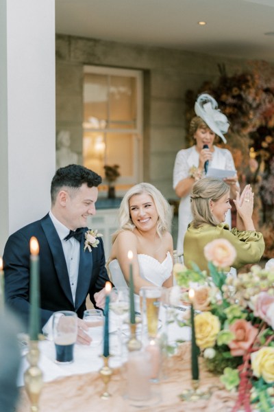 Bride and groom laugh at dining room table during speech by mother