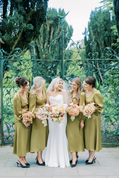 Bride and her bridesmaids stand and pose in front of garden gate they smile