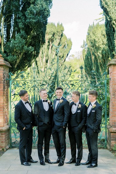 Groom and groomsmen stand in front of gate in garden laughing/smiling