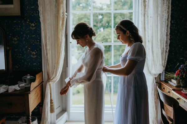 Bridesmaid helps bride with back of dress zip at window