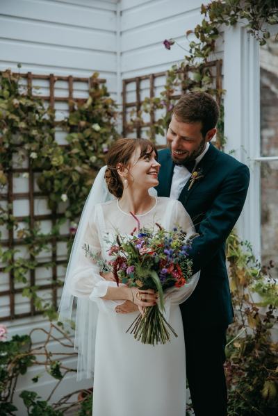 Bride poses in greenhouse with groom hugging her holding bouquet flowers