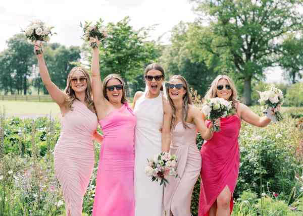 Bride in white and bridesmaids wearing pink dresses holding bouquet hands in the air in garden