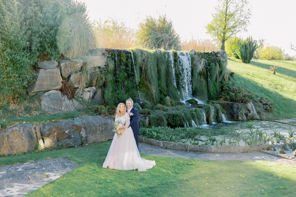 Bride and groom stand in front of scenic waterfall wide shot of couple